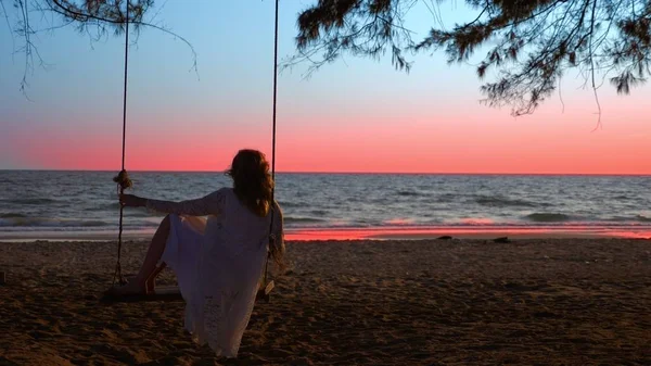 Ein glückliches blondes Mädchen in weißem Kleid und Spitzenumhang schwingt auf einer Seilschaukel am Strand am Meer. — Stockfoto