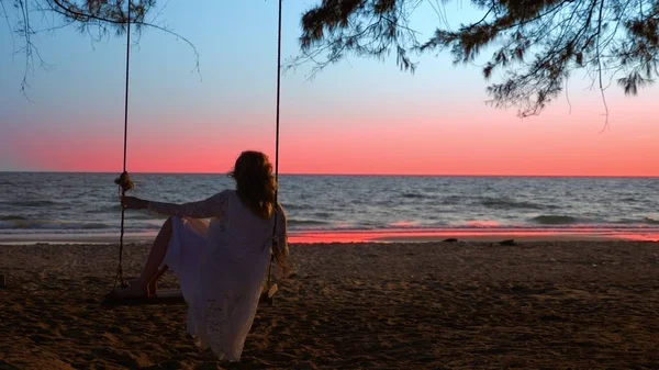 Uma menina loira feliz em um vestido branco e capa de renda está balançando em um balanço de corda, na praia junto ao mar . — Fotografia de Stock