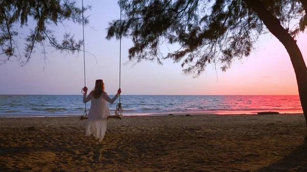 Una chica rubia feliz en un vestido blanco y capa de encaje se balancea en un columpio de cuerda, en la playa junto al mar . — Foto de Stock