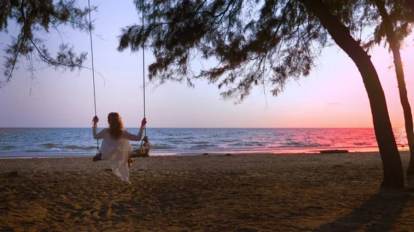 Ein glückliches blondes Mädchen in weißem Kleid und Spitzenumhang schwingt auf einer Seilschaukel am Strand am Meer. — Stockfoto