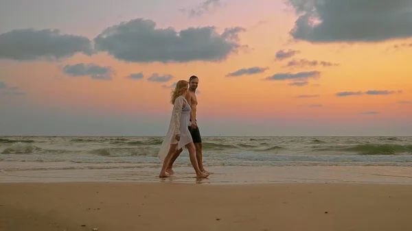 Ρομαντικό ζευγάρι, άνδρας και γυναίκα με τα πόδια κατά μήκος της παραλίας στο ηλιοβασίλεμα, κρατώντας τα χέρια. — Φωτογραφία Αρχείου