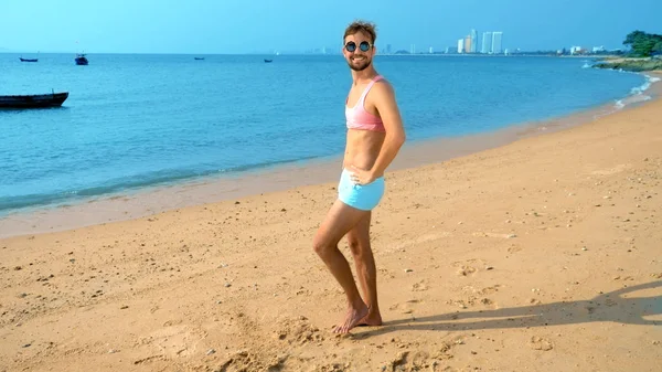 Παιχνιδιάρικο όμορφος άντρας σε μια ροζ μπλούζα και μπλε σορτς χαίρεται στην παραλία. τέρας της θάλασσας. — Φωτογραφία Αρχείου
