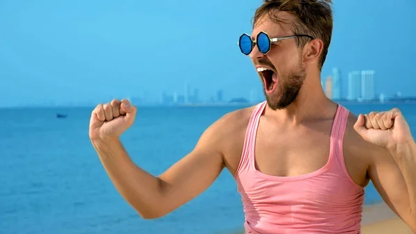 Närbild, lekfull stilig kille i rosa t-shirt på en tropisk strand. han ser på kameran, jublar och gör roliga ansikten — Stockfoto