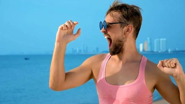 Primo piano, bel ragazzo giocherellone in t-shirt rosa su una spiaggia tropicale. guarda la macchina fotografica, gioisce e fa facce buffe — Foto Stock