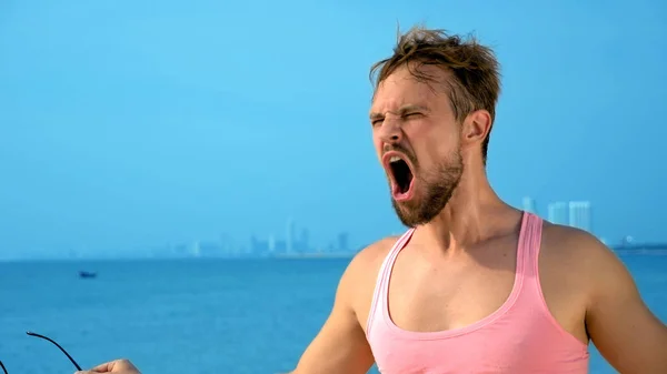 Närbild, lekfull stilig kille i rosa t-shirt på en tropisk strand. han ser på kameran, jublar och gör roliga ansikten — Stockfoto