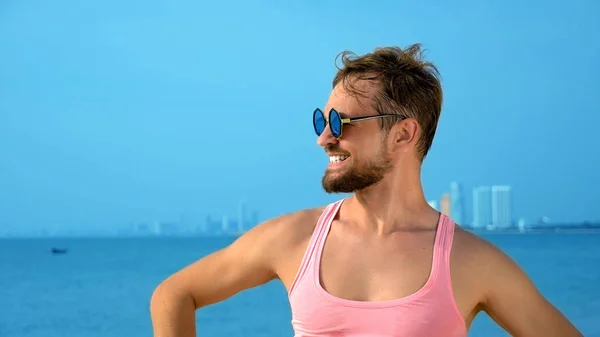 Close-up, Legesyg flot fyr i pink t-shirt på en tropisk strand. han ser på kameraet, glæder sig og gør sjove ansigter - Stock-foto