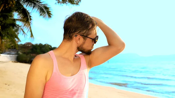 Γκρο πλαν, παιχνιδιάρικο όμορφος άντρας σε ροζ μπλουζάκι σε μια τροπική παραλία. Αυτός κοιτάζει την κάμερα, να χαίρεται και να κάνει αστεία πρόσωπα — Φωτογραφία Αρχείου