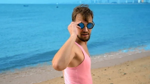 Närbild, lekfull stilig kille i rosa t-shirt på en tropisk strand. han ser på kameran, jublar och gör roliga ansikten — Stockfoto