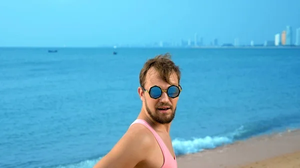 Närbild, lekfull stilig kille i rosa t-shirt på en tropisk strand. han ser på kameran, jublar och gör roliga ansikten — Stockfoto