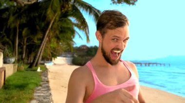Yakın çekim, tropikal bir plajda pembe tişört oynak yakışıklı adam. o da kameraya baktı, sevinir ve komik yüz yapıyor