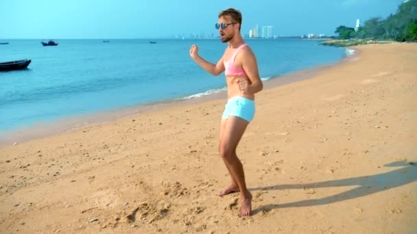 Der verspielte, gutaussehende Kerl in rosa T-Shirt und blauer Shorts jubelt am Strand. Freak auf dem Meer. — Stockvideo