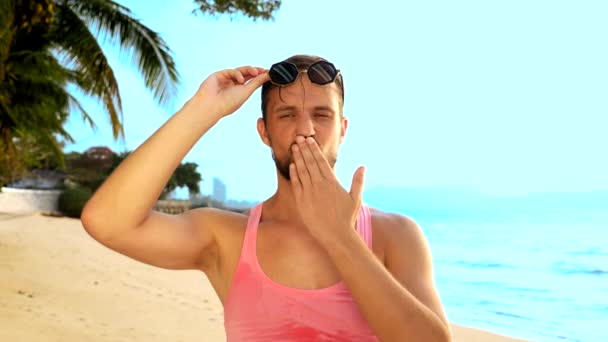 Närbild, lekfull stilig kille i rosa t-shirt på en tropisk strand. han ser på kameran, jublar och gör roliga ansikten — Stockvideo