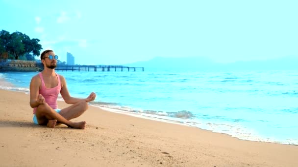 Playful handsome man in pink t-shirt and blue shorts meditates while sitting by the sea — Stock Video