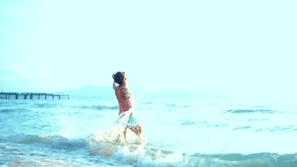 Um cara bonito brincalhão com uma camiseta rosa e shorts azuis corre alegremente para o mar. aberração no mar — Vídeo de Stock