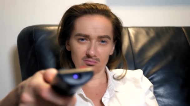 Portrait d'une féministe androgyne avec une moustache peinte sur son visage assis sur le canapé et commutant les chaînes de télévision du panneau de commande — Video