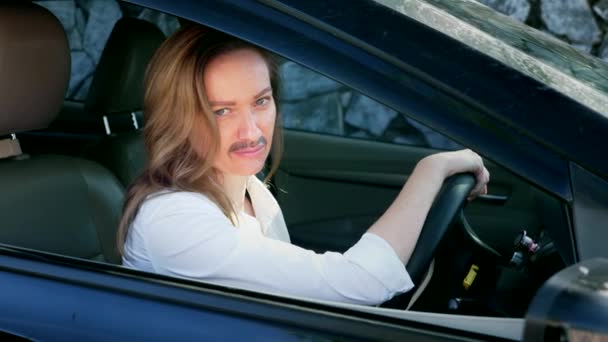 Un retrato de una feminista andrógina con un bigote pintado en la cara sentada al volante de un coche. mujer mira a la cámara desde la ventana del coche . — Vídeos de Stock