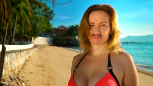 Das Konzept der seltsamen Abenteuer der Menschen. eine schöne Frau im rosafarbenen Bikini am Strand, der Kamera zugewandt, und ihr Schnurrbart ist auf ihrem Gesicht sichtbar. — Stockvideo