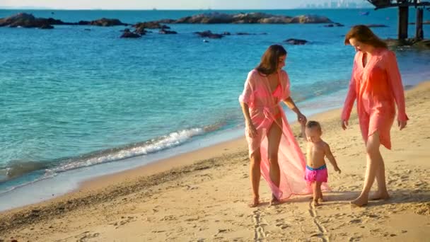 Le concept des étranges aventures des gens. Joyeux couple lesbien avec bébé marchant sur la belle plage tropicale. une des femmes avec une moustache sur le visage comme symbole d'émancipation . — Video