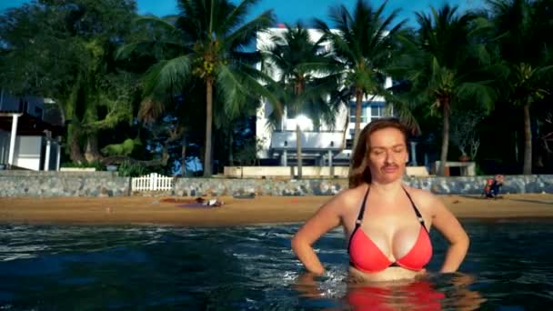 El concepto de las extrañas aventuras de la gente. Hermosa mujer femenina en bikini rosa con bigote en la cara como símbolo de emancipación, salpicando en el mar contra una playa tropical — Vídeo de stock