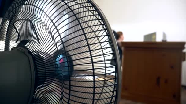 Primer plano de una parte de un ventilador de oficina, sobre el fondo de un escritorio borroso, en el que una persona trabaja en un ordenador — Vídeo de stock