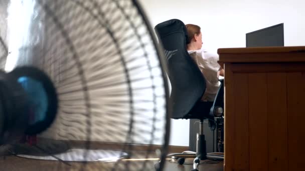 Närbild av en del av en office-fläkt, i bakgrunden suddig skrivbord, där en person arbetar på en dator — Stockvideo