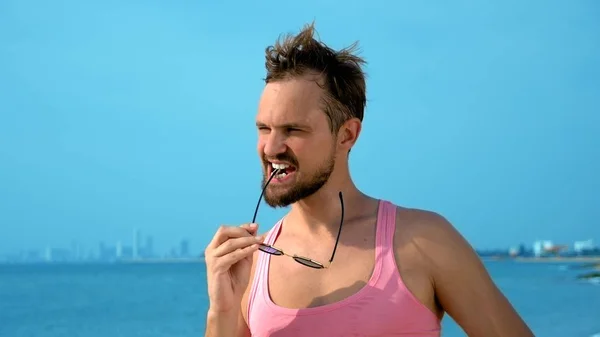 Lekfull stilig kille i en rosa t-shirt och blå shorts jublar på stranden. Freak på havet. — Stockfoto