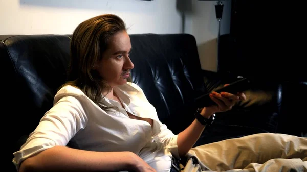 Retrato de una feminista andrógina con un bigote pintado en la cara sentada en el sofá y cambiando los canales de televisión desde el panel de control — Foto de Stock