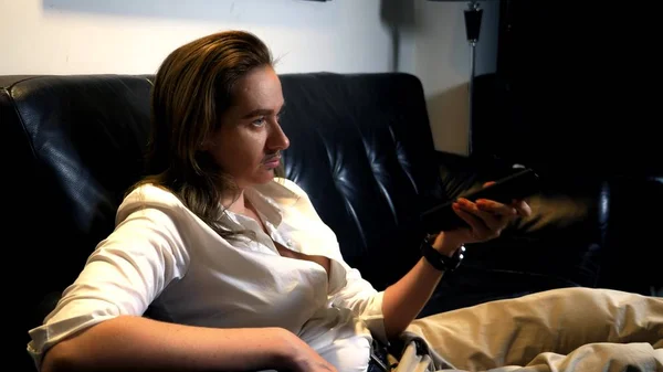 portrait of an androgynous feminist with a painted mustache on her face sitting on the sofa and switching TV channels from the control panel