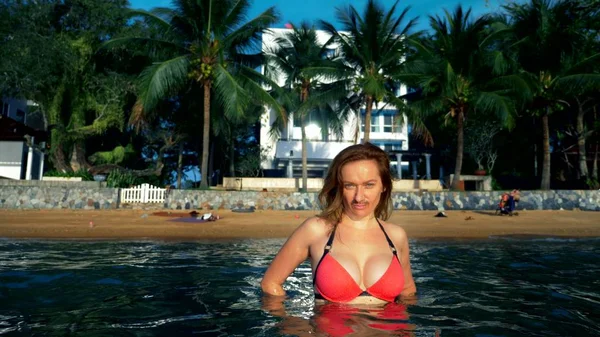 El concepto de las extrañas aventuras de la gente. Hermosa mujer femenina en bikini rosa con bigote en la cara como símbolo de emancipación, salpicando en el mar contra una playa tropical — Foto de Stock