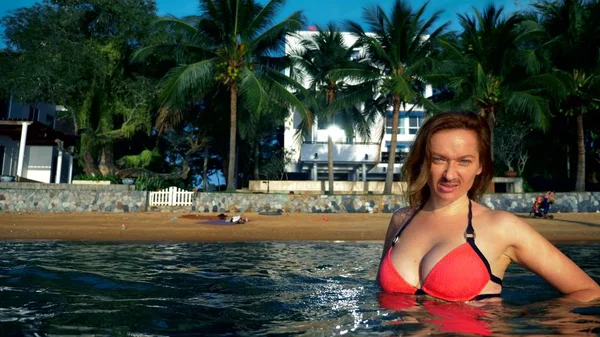 El concepto de las extrañas aventuras de la gente. Hermosa mujer femenina en bikini rosa con bigote en la cara como símbolo de emancipación, salpicando en el mar contra una playa tropical — Foto de Stock