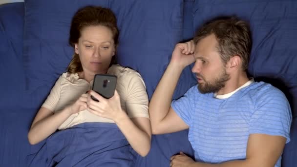 セックスの問題の概念。ベッドのカップル。彼の妻は関係の彼を無視して携帯電話を使用している間、夫は動揺 — ストック動画