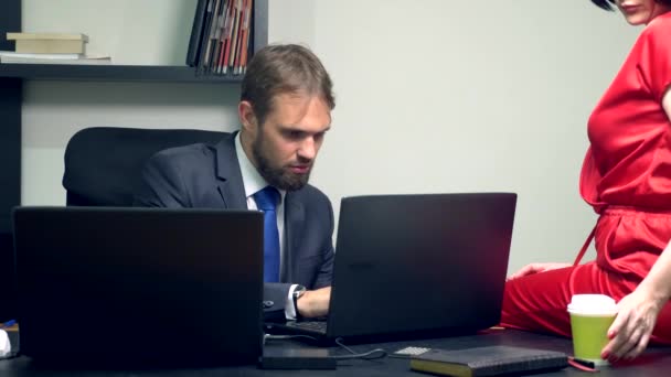 Biuro flirt. Atrakcyjna kobieta w czerwonym kombinezon siedzi na biurku jej kolega i zmyw dotyka jego szklankę kawy. — Wideo stockowe