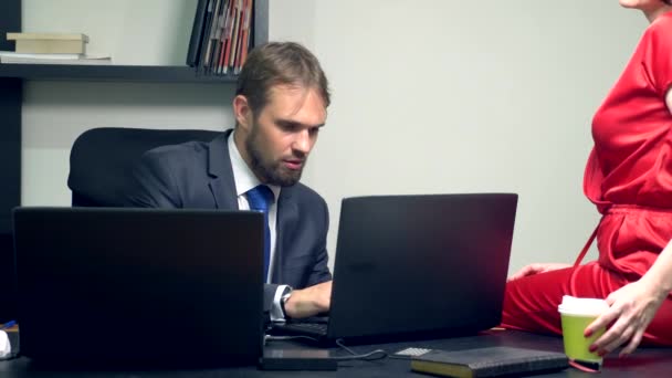 Biuro flirt. Atrakcyjna kobieta w czerwonym kombinezon siedzi na biurku jej kolega i zmyw dotyka jego szklankę kawy. — Wideo stockowe