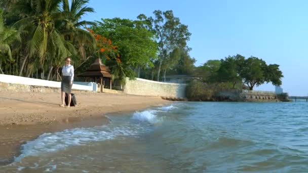 Geschäftsfrau in Bürokleidung mit Koffer läuft barfuß an einem weißen Sandstrand vor der Kulisse von Palmen und einem Luxus-Resort entlang. freiberufliches, langersehntes Freizeitkonzept — Stockvideo