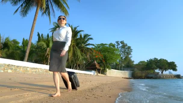 Mulher de negócios em roupas de escritório com uma mala está andando descalça ao longo de uma praia de areia branca contra o pano de fundo de palmeiras e um resort de luxo. freelance, conceito de lazer tão esperado — Vídeo de Stock