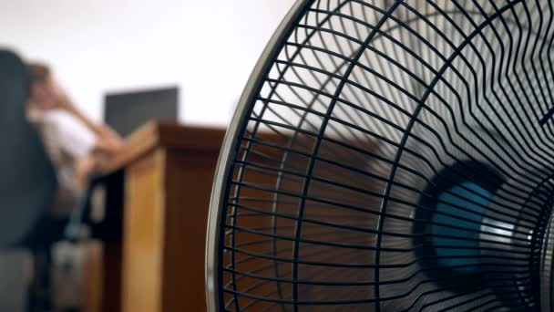 Primo piano di una parte di un ventilatore da ufficio, sullo sfondo di una scrivania sfocata, presso la quale una persona lavora in un computer — Video Stock