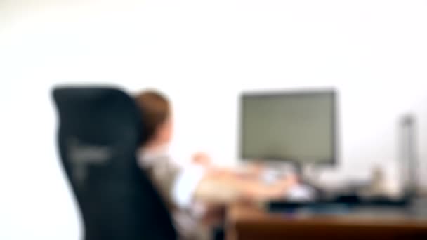 Крупным планом части офисного вентилятора, на фоне размытого письменного стола, за которым человек работает за компьютером — стоковое видео