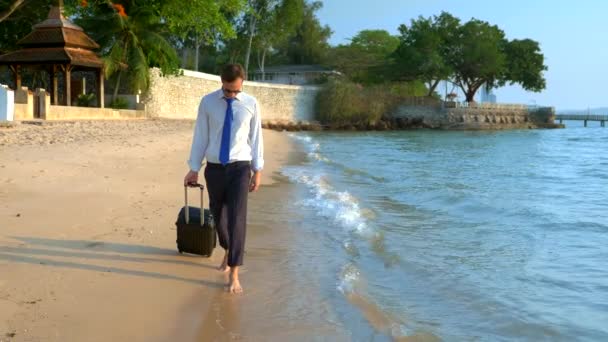 Bello uomo d'affari in occhiali da sole con una valigia va a piedi nudi sulla spiaggia di sabbia bianca sullo sfondo di palme e un resort di lusso. freelance, concetto di svago tanto atteso — Video Stock