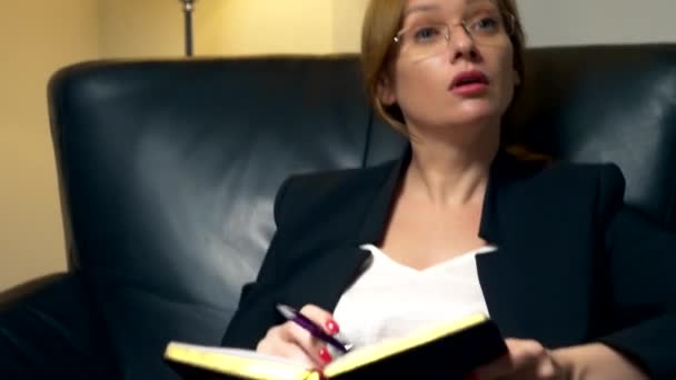 Gros plan. une femme parle à quelqu'un et prend des notes dans un carnet alors qu'elle est assise le soir sur une chaise en cuir noir au bureau. Le concept de consultation, de négociation . — Video