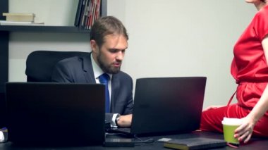 Ofis flört. Kırmızı bir tulum içinde çekici bir kadın onun meslektaşı masasında oturuyor ve sensually kahve onun bardak dokunur.