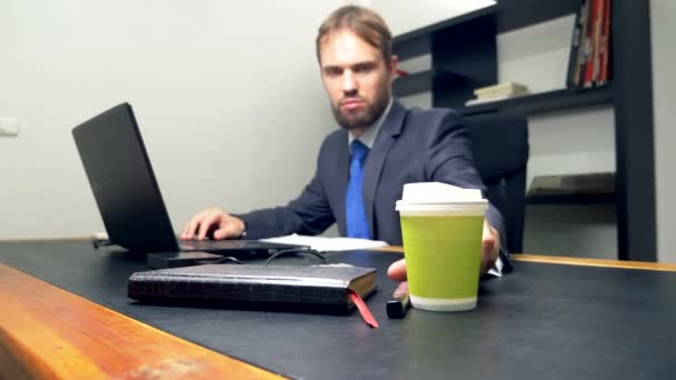 Un hombre de traje trabaja en un escritorio de la oficina en un portátil y bebe café de una taza desechable. espacio de copia — Vídeo de stock