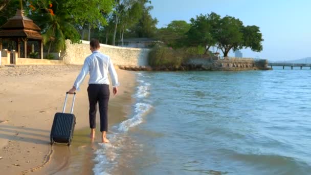 Красивий бізнесмен в сонцезахисних окулярах з валізою йде босоніж на білому піщаному пляжі на тлі пальмових дерев і розкішного курорту. вільний, довгоочікувана концепція дозвілля — стокове відео