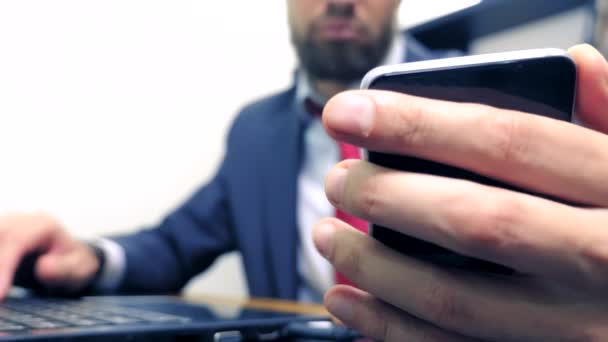 Närbild. Affärs man använder mobil smartphone och leende, medan du arbetar på en bärbar dator på kontoret. — Stockvideo