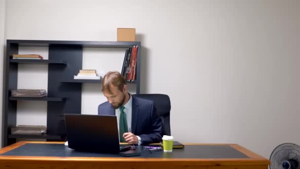 Seorang pria dalam setelan jas bekerja di sebuah meja di kantor pada laptop dan minum kopi dari cangkir sekali pakai. menyalin ruang — Stok Video