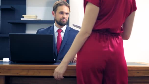 Office flirta. Attraktiv kvinna i röda overaller med en djup urringning flirta vid bordet med sin kollega. man tittar på bröstet av en kvinna på kontoret — Stockvideo