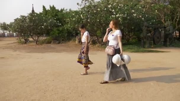 Dos niñas, madre e hija, turistas pasan por el territorio de un templo budista de madera en faldas largas. con cascos en la mano . — Vídeos de Stock