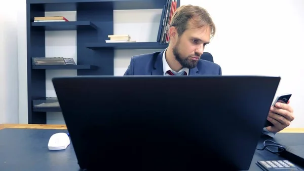 Gros plan. Homme d'affaires utilisant un smartphone mobile et souriant, tout en travaillant sur un ordinateur portable dans le bureau . — Photo
