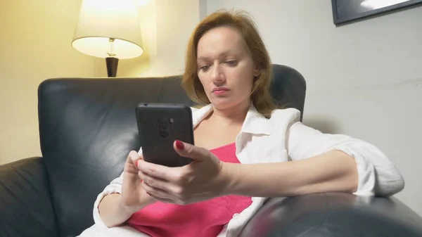 Una mujer está utilizando un teléfono inteligente en casa, sentado en una silla de cuero grande por la noche, en el fondo una lámpara de pie — Foto de Stock