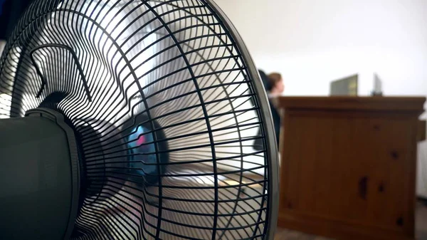 close-up of part of an office fan, on the background of a blurred writing desk, at which a person works at a computer