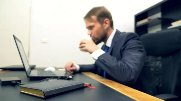 Un hombre de traje trabaja en un escritorio de la oficina en un portátil y bebe café de una taza desechable. espacio de copia — Foto de Stock